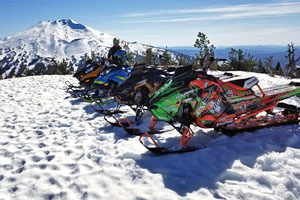 Row of snowmobiles on snow-covered peak with Mt. Bachelor in the background
