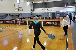 Pickleball courts set up on a hardwood floor with several players engaged in doubles play.