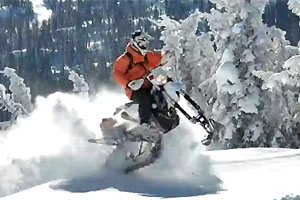 Snowbike catching some air off a snowfield with snow-covered trees in the background.