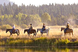 Four people on horseback riding along a body of water with pine forests in the background.