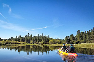 Couple canoeing on the Deschutes River with a mirror surface and pine forest in the background.